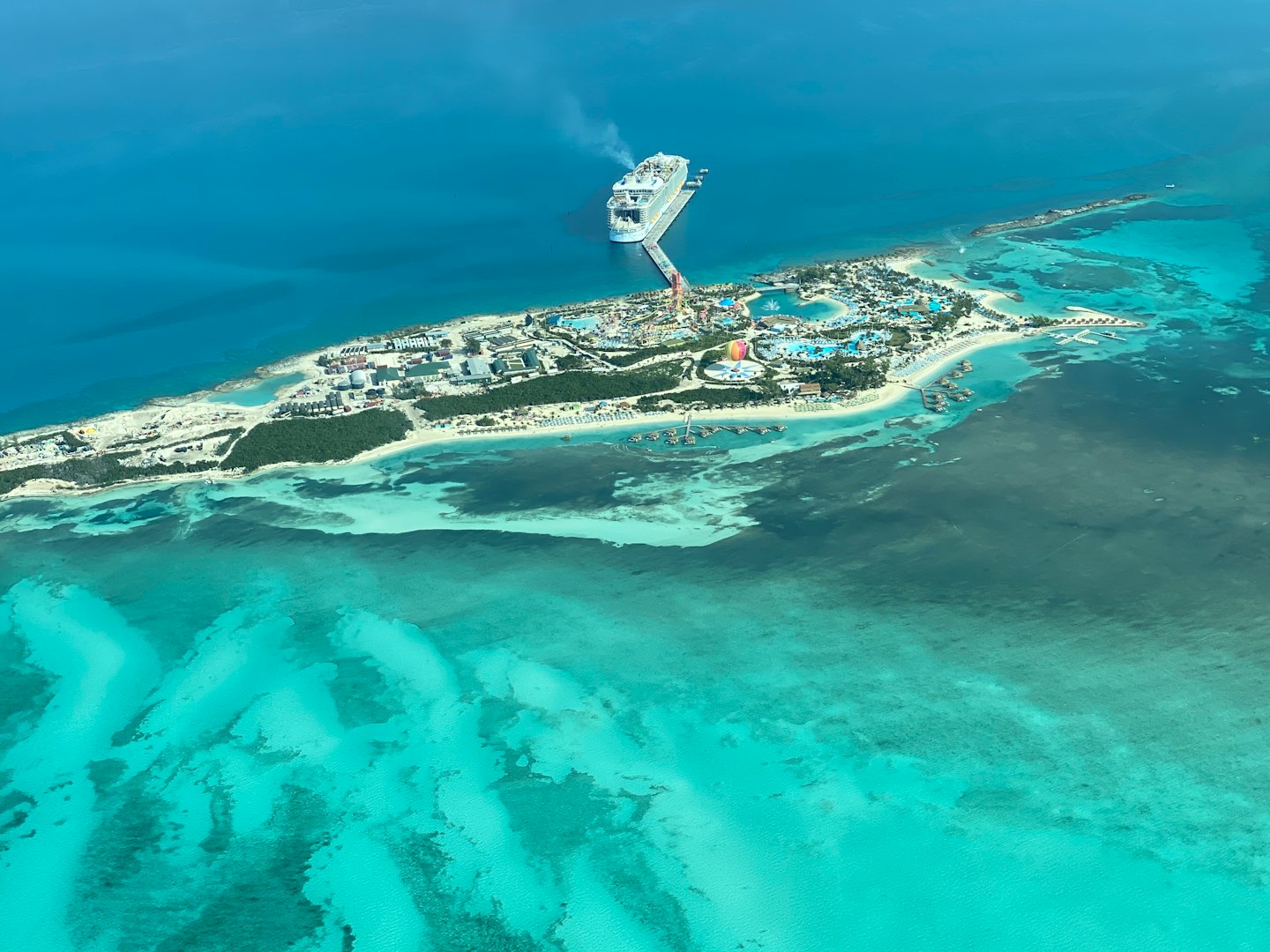 Weißes Kreuzfahrtschiff, das in einem tropischen Inselresort mit Pools und Bungalows angedockt ist.
